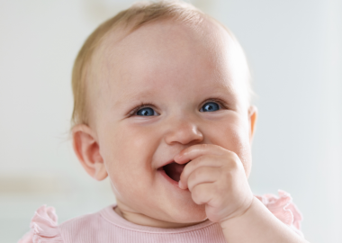 Close-up of a smiling baby with blue eyes, wearing a pink outfit and finger in mouth